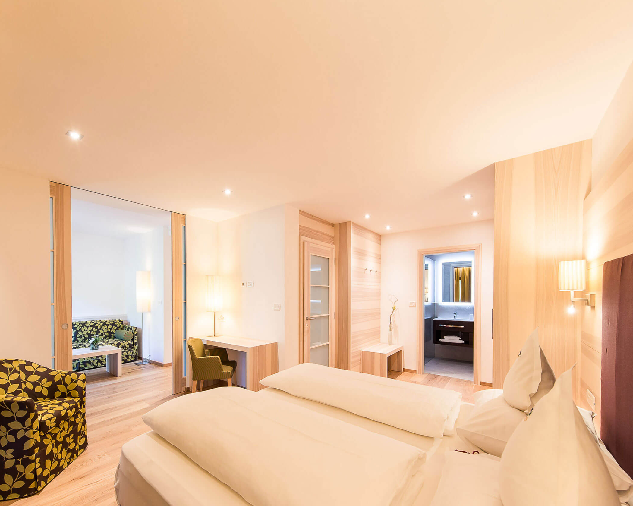 Bright, modern hotel room with double bed, armchair, desk, and view into sitting area and bathroom. - Hotel Lagrein