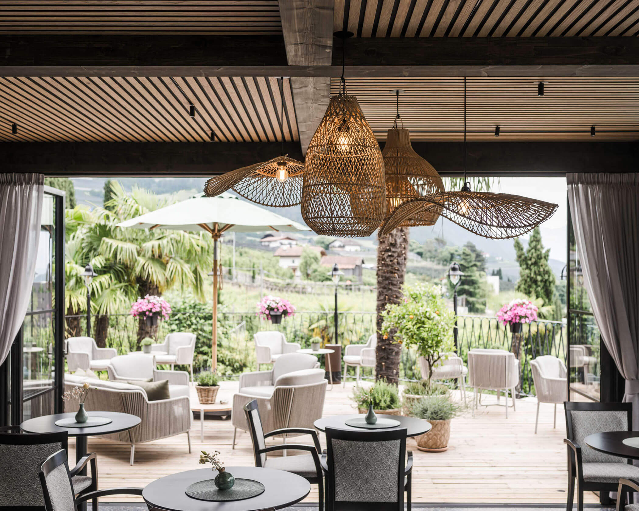 Chic outdoor patio with wicker lights, modern seating, and lush greenery overlooking scenic countryside. - Hotel Lagrein
