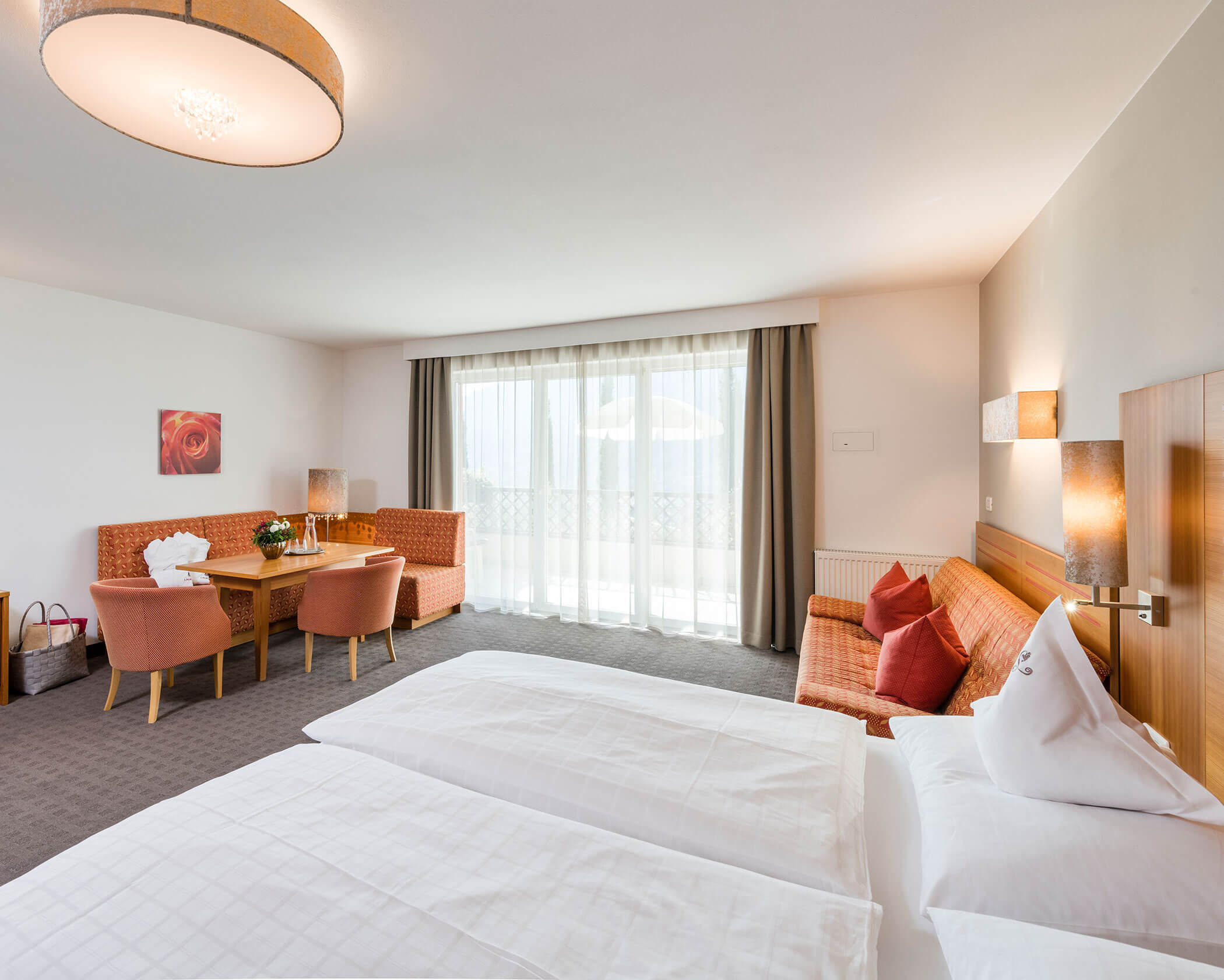 Bright hotel room with a double bed, orange chairs, sofa, table, and large window with sheer curtains. - Hotel Lagrein