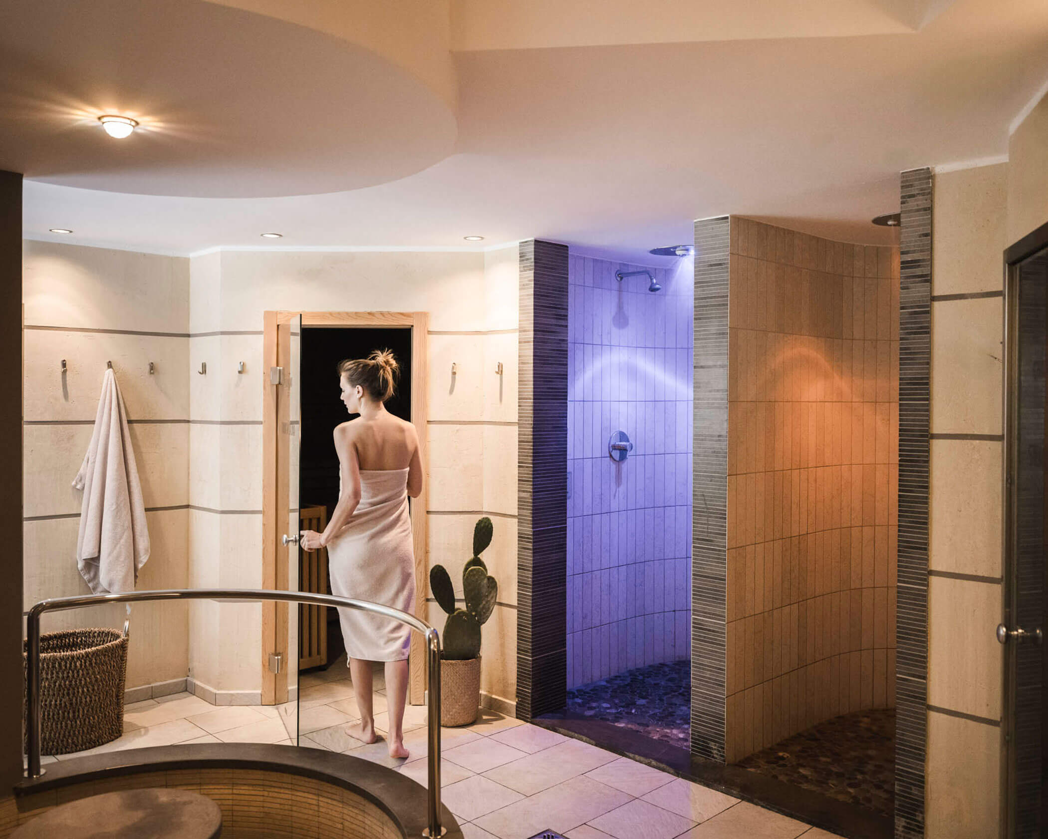 A woman in a towel walks toward a wooden sauna in a modern spa with showers and a hot tub. - Hotel Lagrein