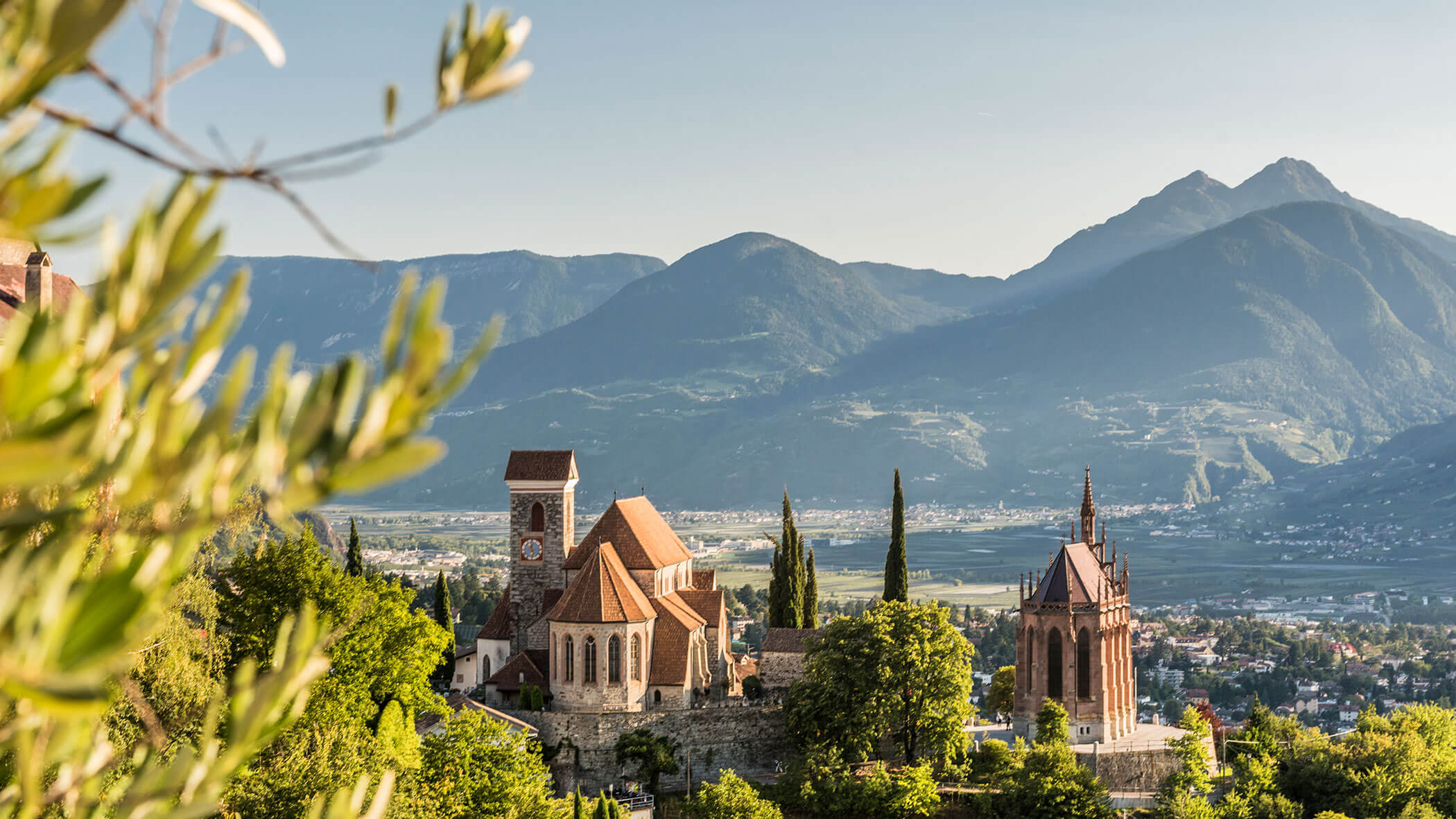 A picturesque village with historic churches, trees, and mountains in the background under a clear sky. - Hotel Lagrein