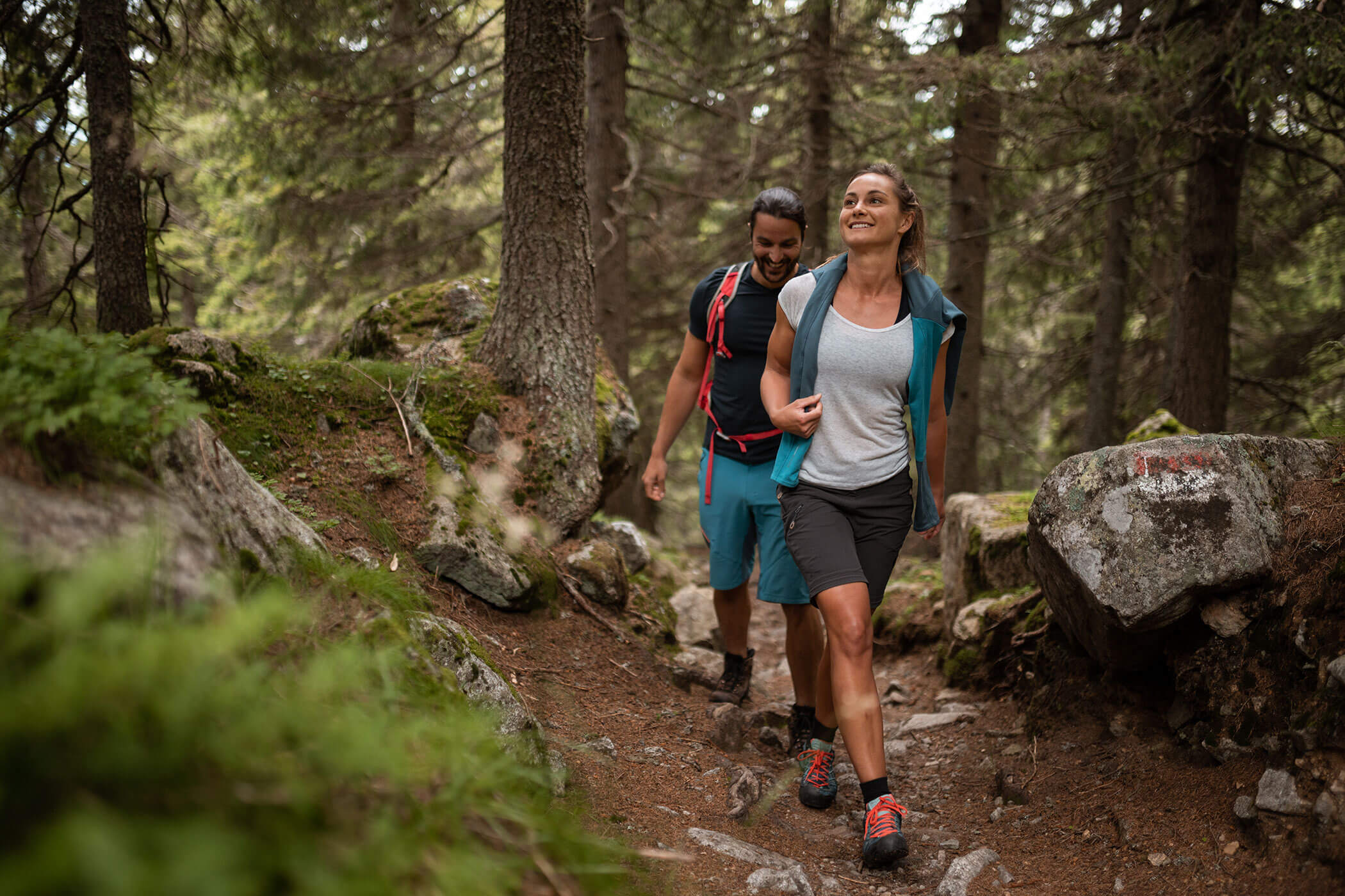 Two people hike on a rocky forest trail, surrounded by trees and greenery, smiling and enjoying nature. - Hotel Lagrein