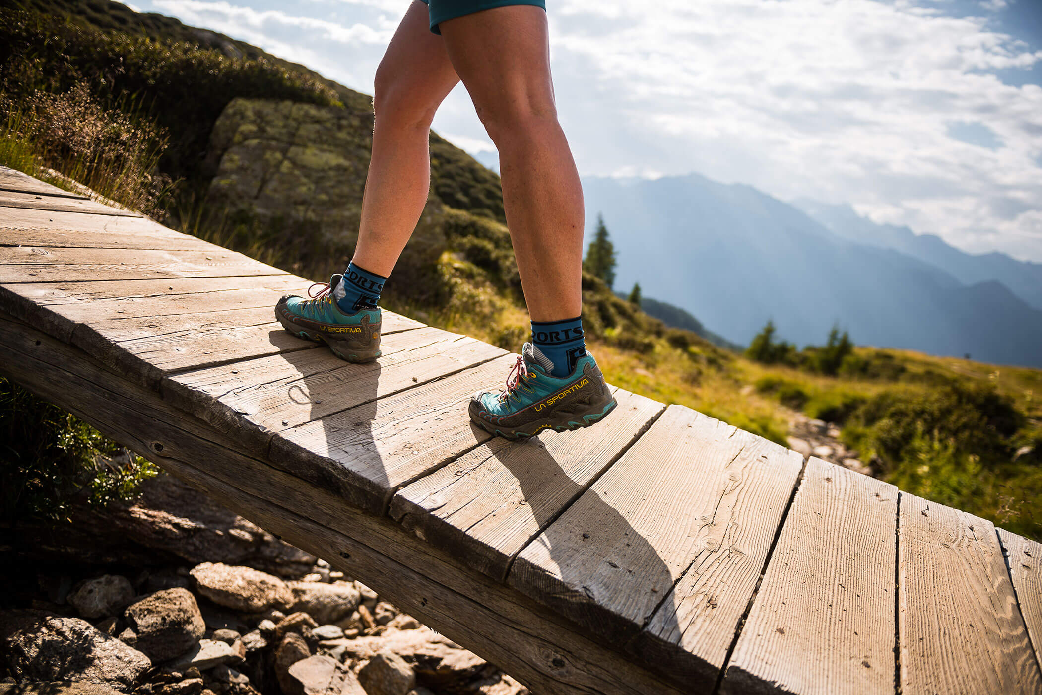 Person hiking up a wooden path in the mountains on a sunny day, wearing teal trail shoes and blue socks. - Hotel Lagrein