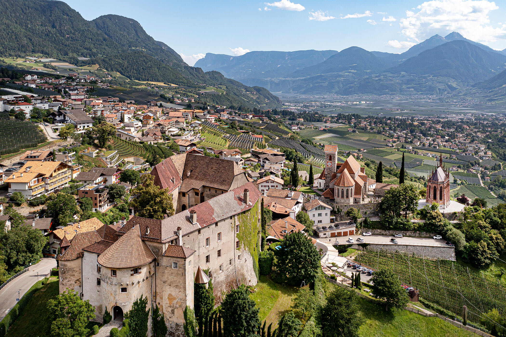 A hilltop village with historic buildings, vineyards, and mountains in the background under a blue sky. - Hotel Lagrein
