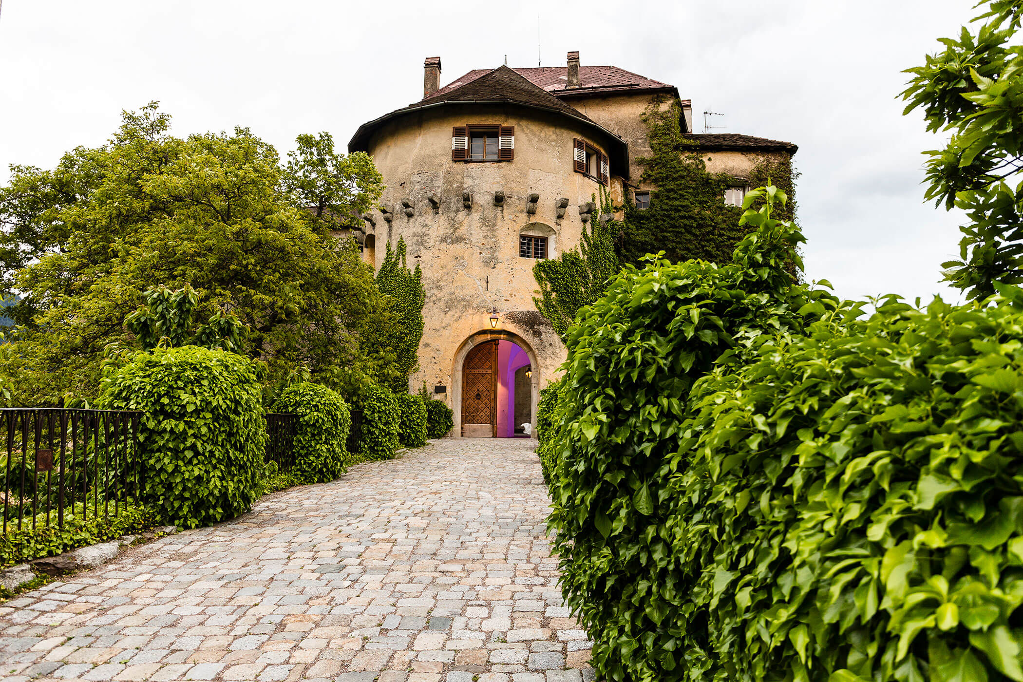 A stone path leads to an old, ivy-covered castle entrance surrounded by lush green bushes. - Hotel Lagrein