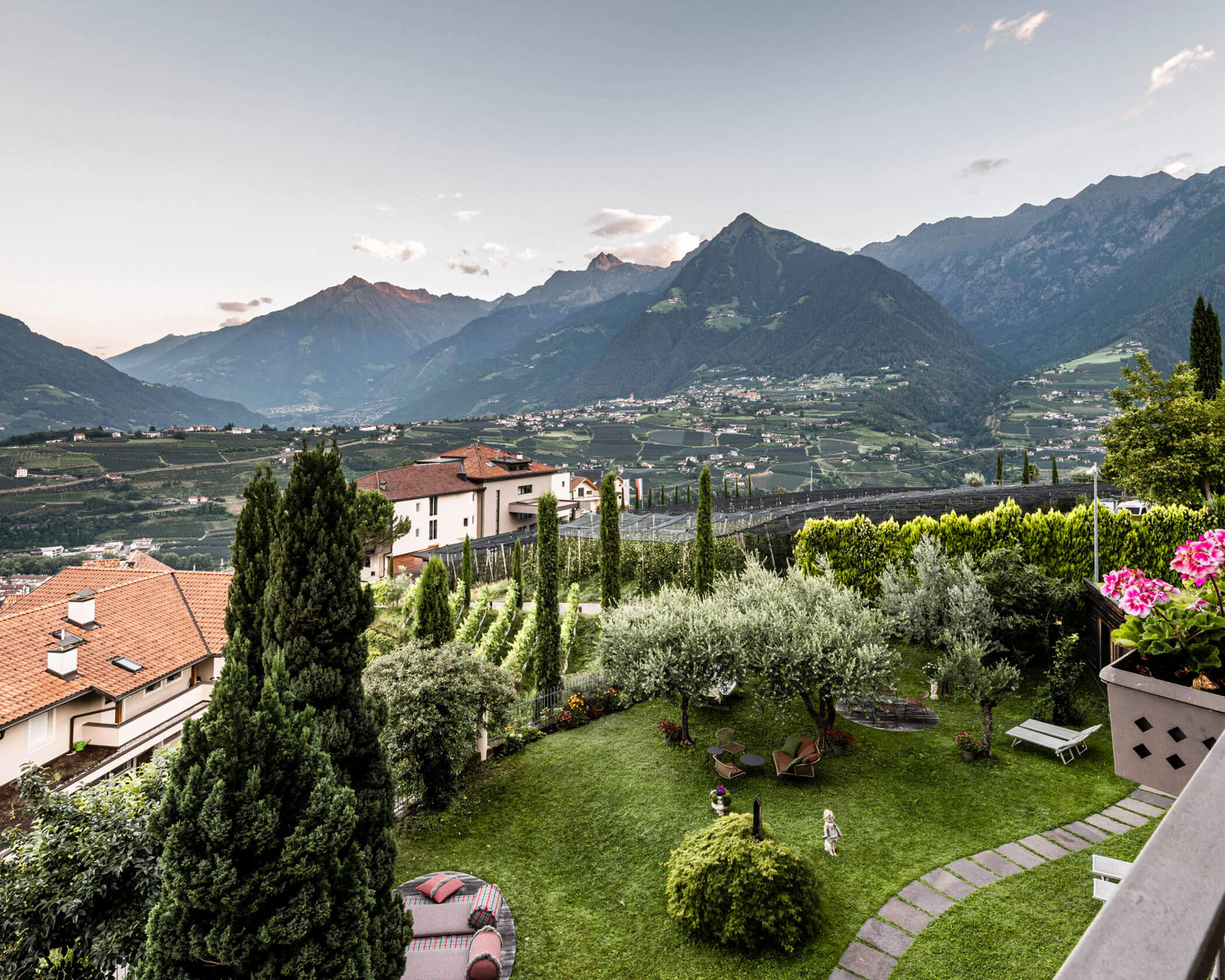 Scenic mountain village with green gardens, houses, and distant peaks under a cloudy sky at sunset. - Hotel Lagrein
