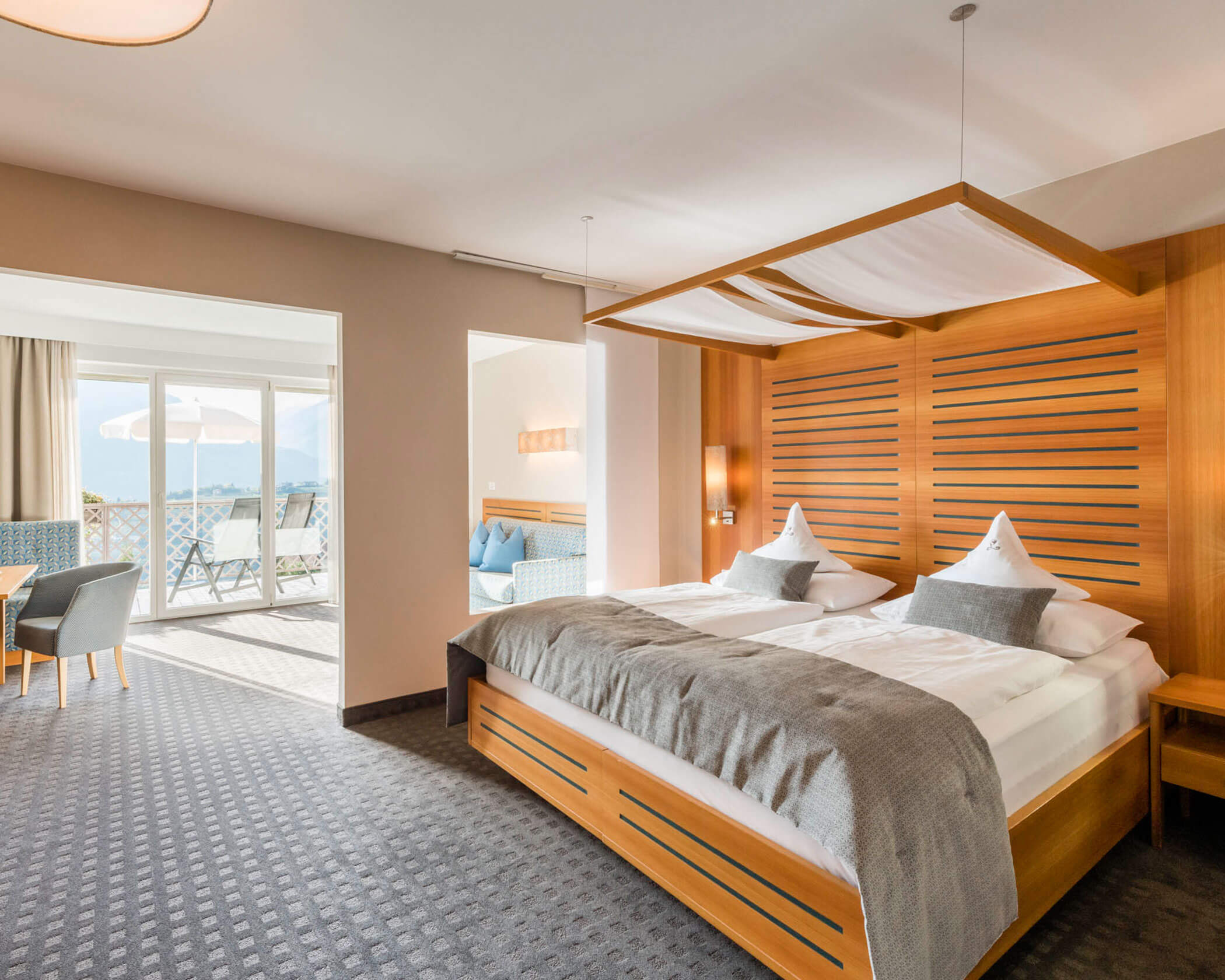 Bright hotel room with a large bed, wooden accents, seating area, and a balcony with outdoor chairs. - Hotel Lagrein