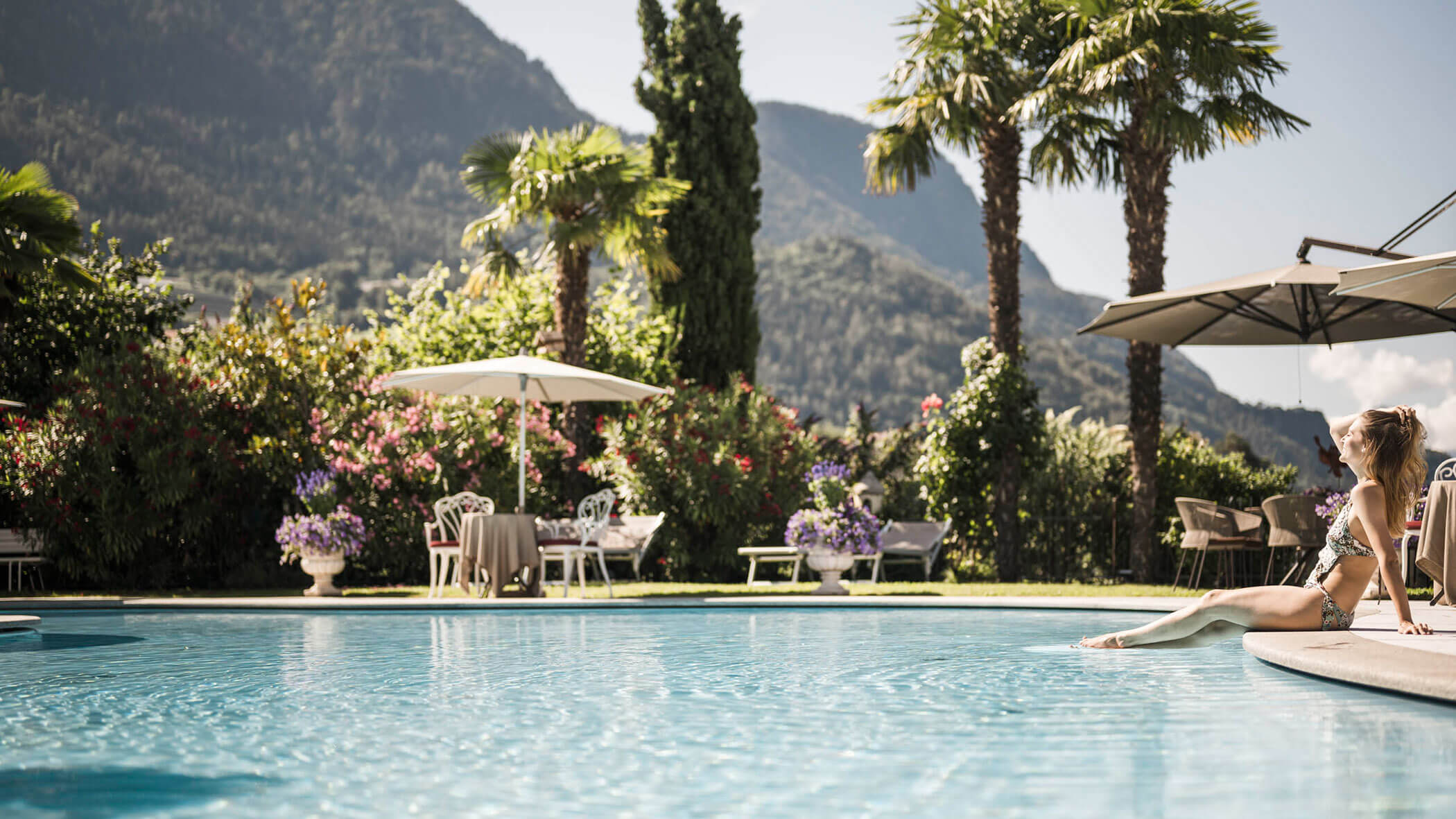 Woman relaxing at the edge of a pool with palm trees, mountains, and patio tables in the background. - Hotel Lagrein