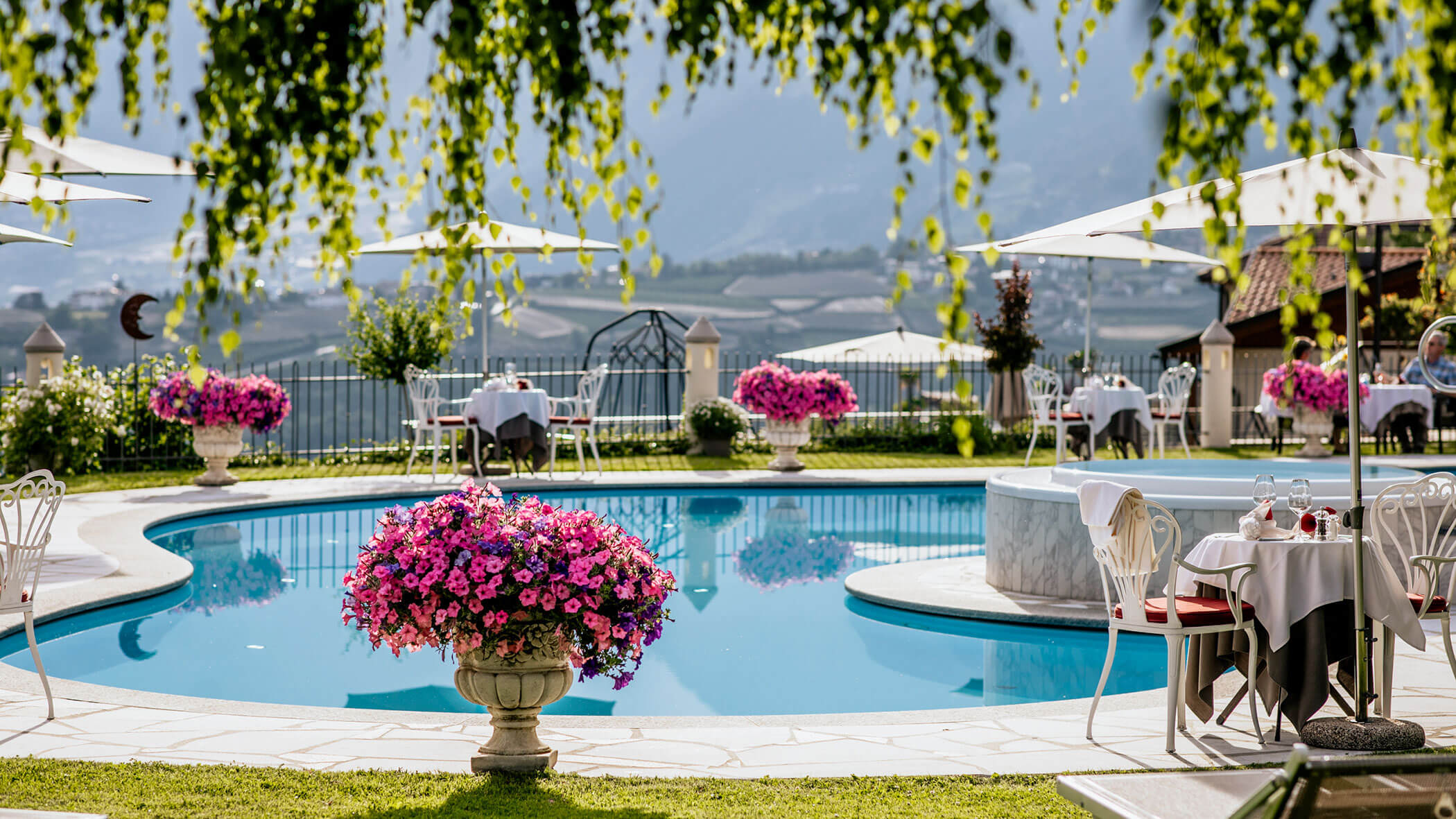 Outdoor pool area with flower pots, tables set for dining, and sun umbrellas, overlooking a scenic landscape. - Hotel Lagrein