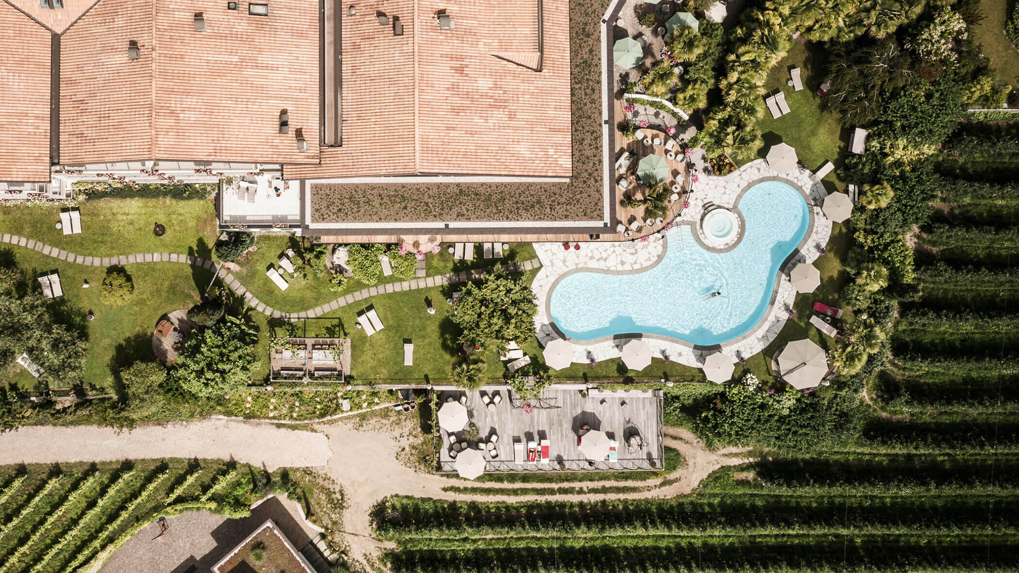Aerial view of a pool, garden, and building surrounded by greenery and lounge chairs. - Hotel Lagrein