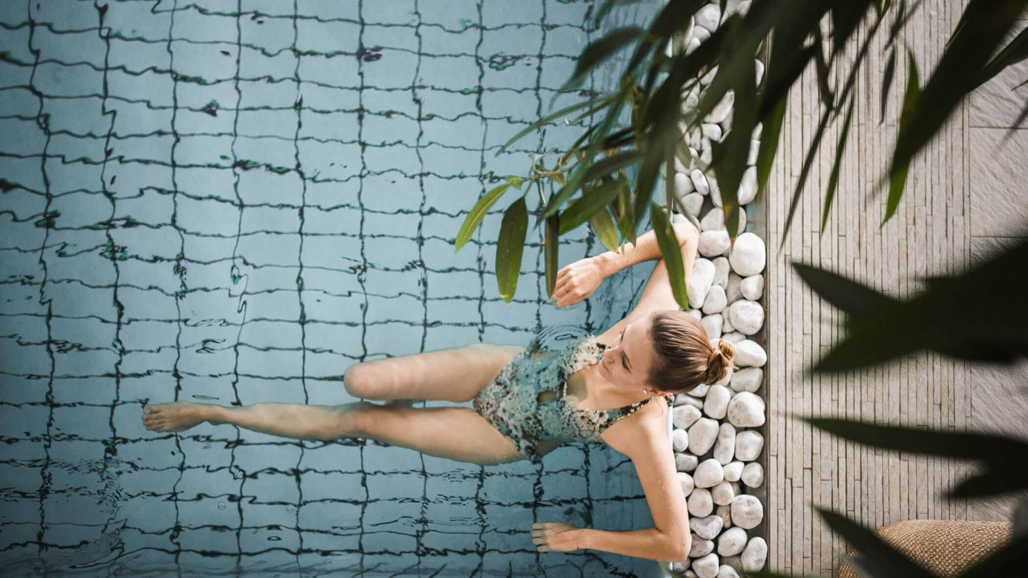 Woman in a patterned swimsuit relaxing in a pool near white stones and green plants, viewed from above. - Hotel Lagrein