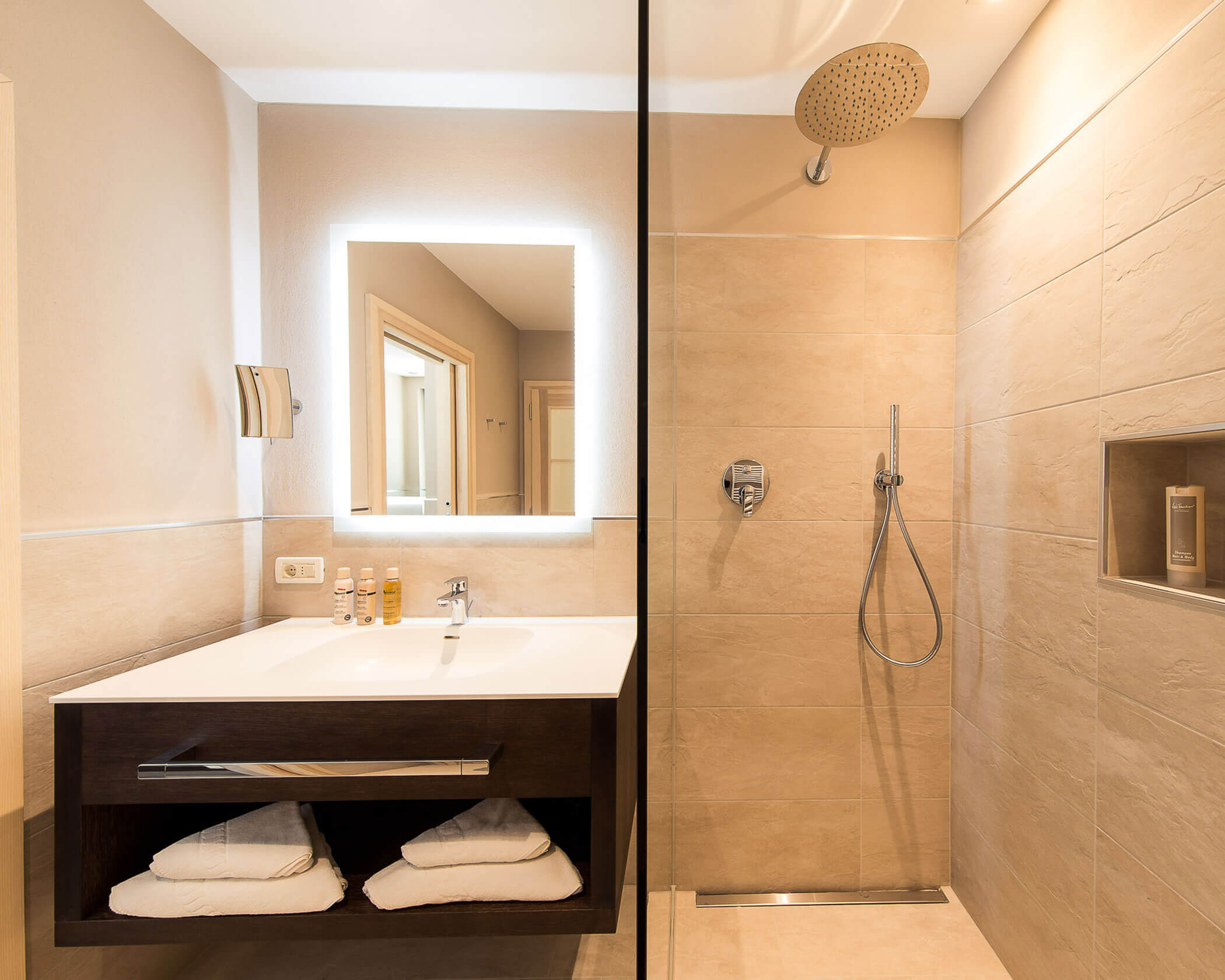 Modern bathroom with a lit mirror, sink, towels, and a walk-in shower with a rainfall showerhead. - Hotel Lagrein