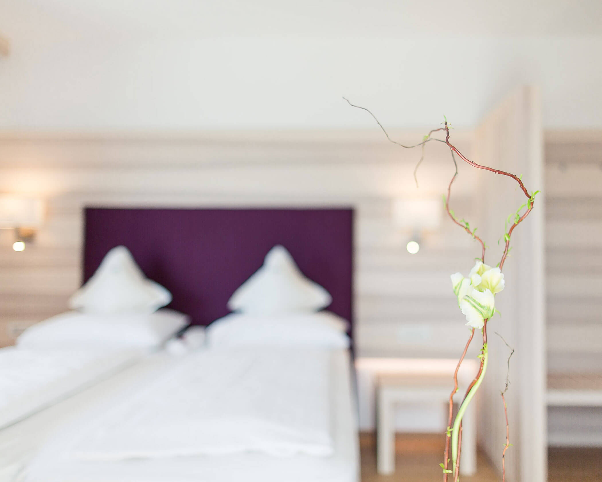 A blurred hotel room with two beds, white linens, and a flower in focus in the foreground. - Hotel Lagrein