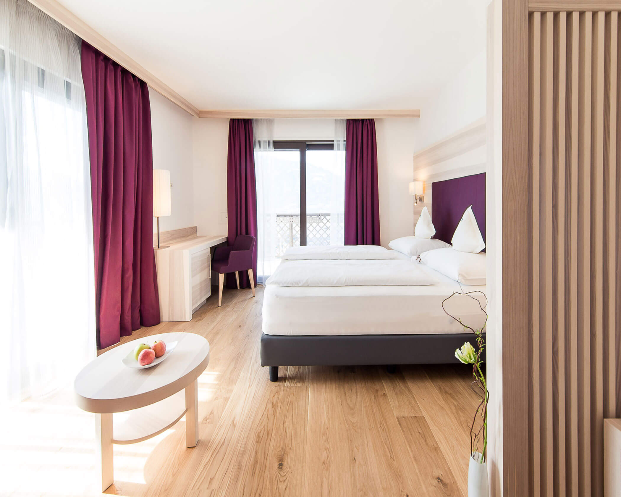 Bright hotel room with two beds, purple curtains, a desk, and a balcony door letting in natural light. - Hotel Lagrein