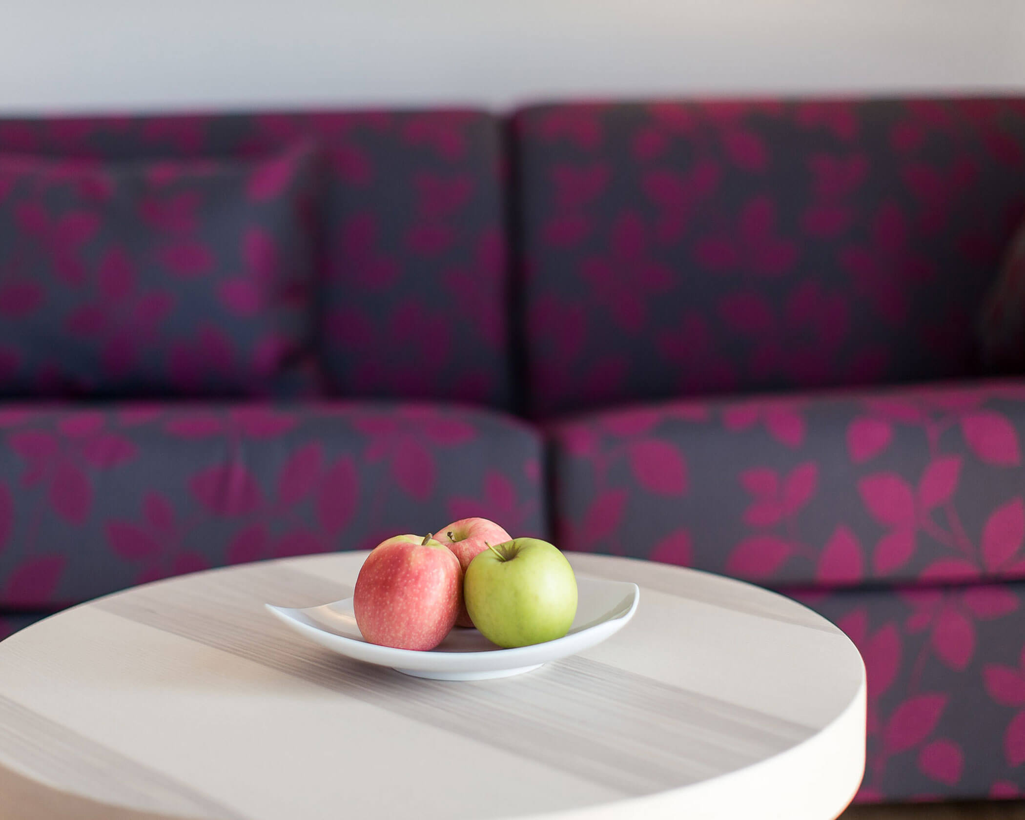 Three apples—two red and one green—on a white plate atop a round table, with a purple sofa in the background. - Hotel Lagrein