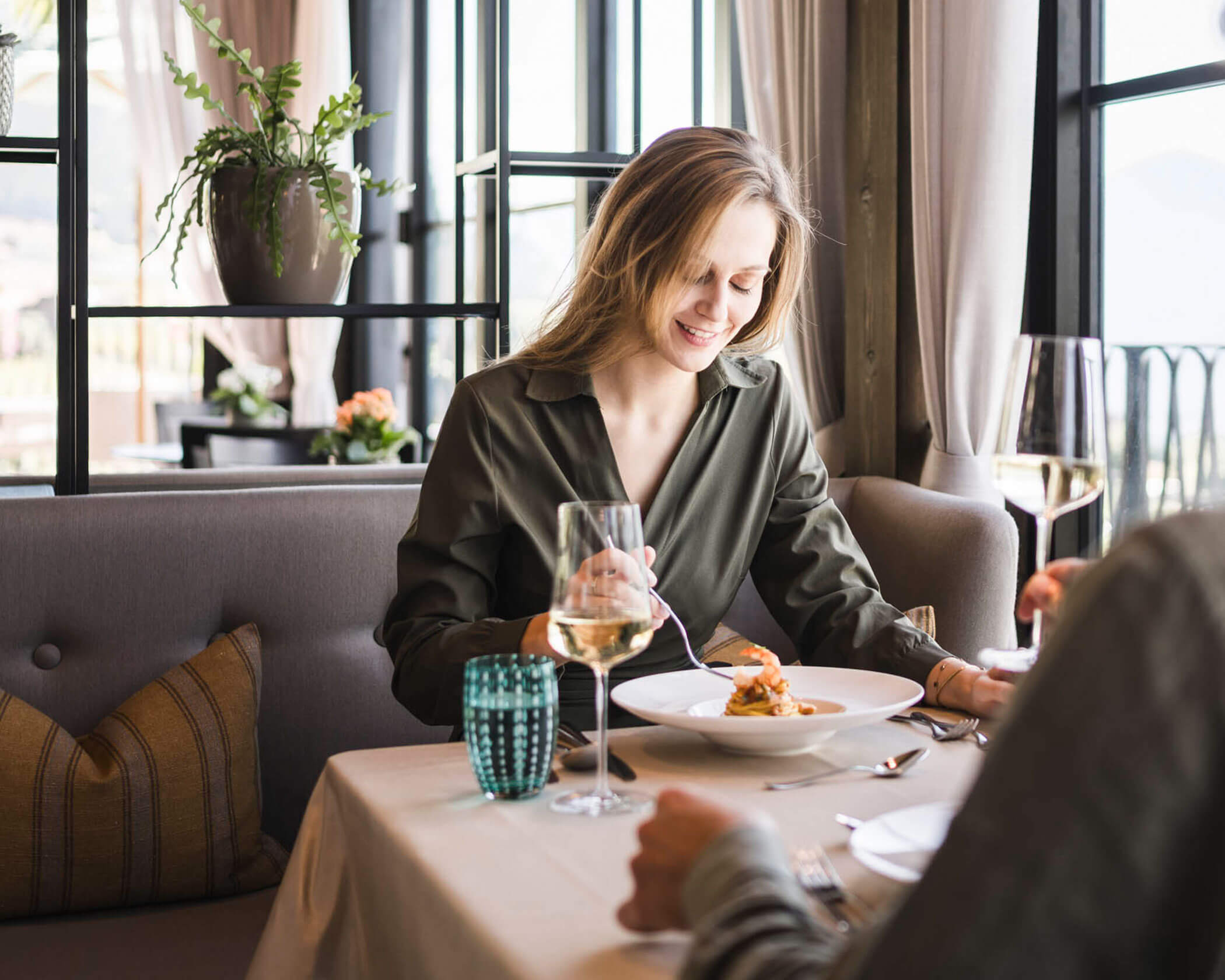 Woman smiling and eating at a restaurant table with wine and water glasses, sitting across from another person. - Hotel Lagrein