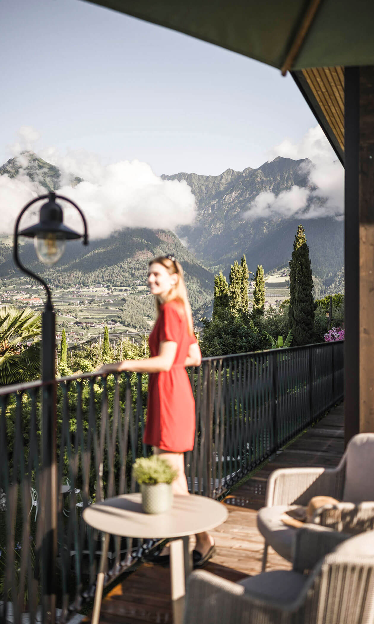 Woman in a red dress stands on a balcony overlooking mountains and lush greenery on a sunny day. - Hotel Lagrein