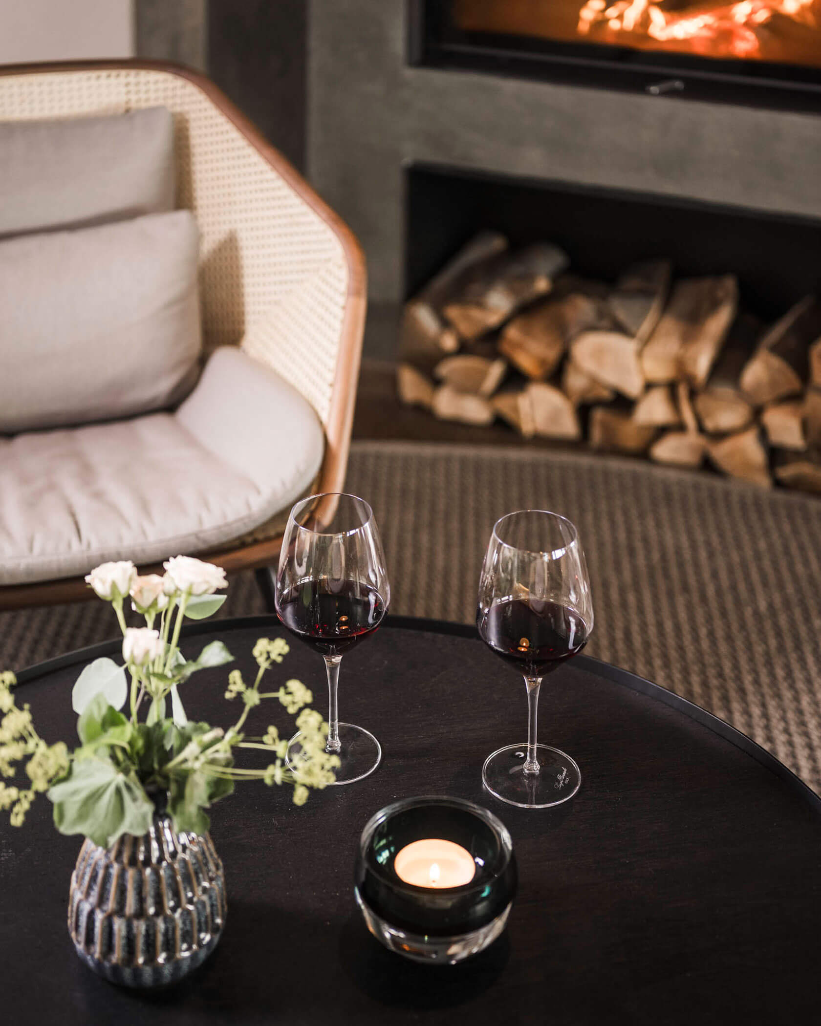 Two glasses of red wine on a table with flowers and a candle, cozy chair and firewood in the background. - Hotel Lagrein