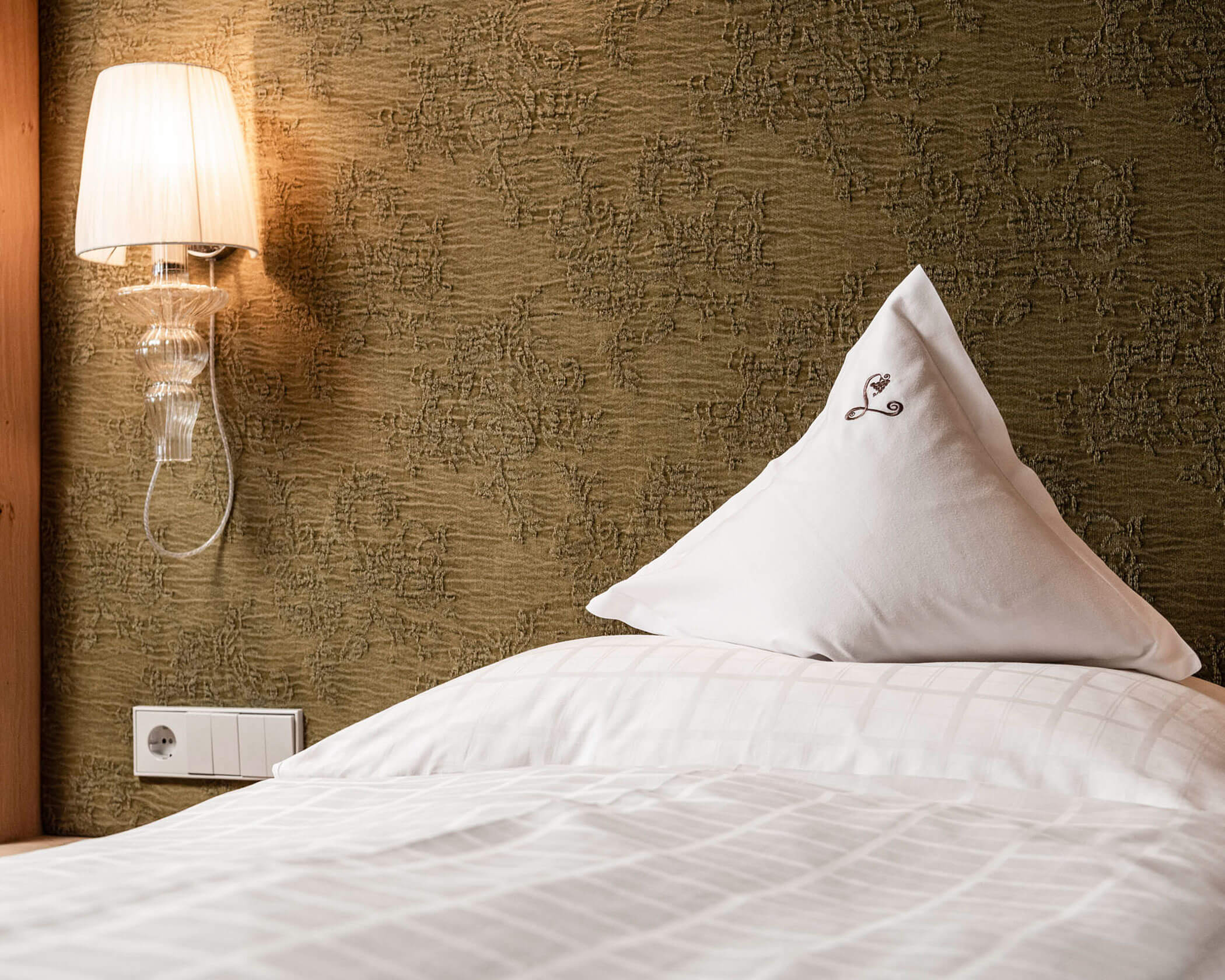 A neatly made bed with a plaid white pillow under a wall lamp on a textured brown wall. - Hotel Lagrein