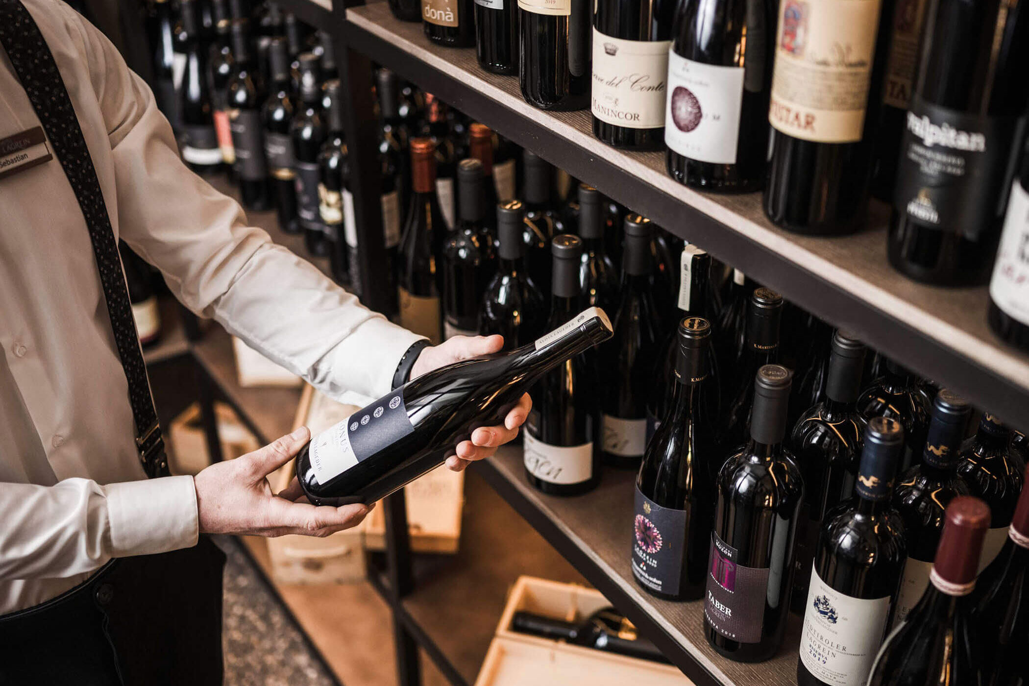 A person holding a wine bottle in front of shelves filled with various bottles of wine. - Hotel Lagrein