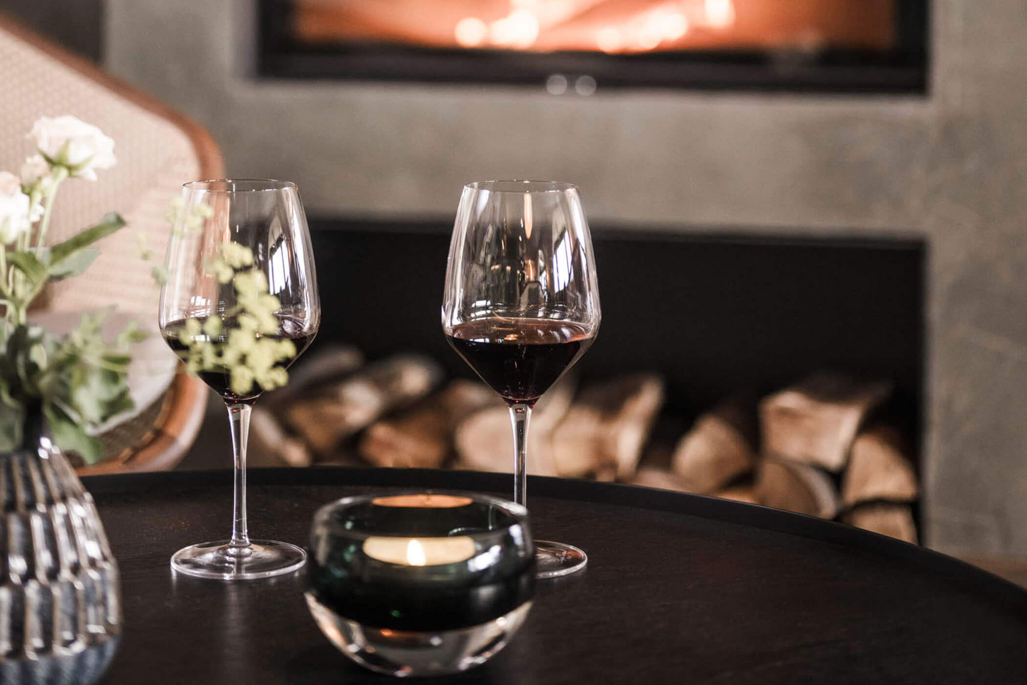 Two glasses of red wine on a table with a candle, flowers, and a fireplace with stacked logs in the background. - Hotel Lagrein