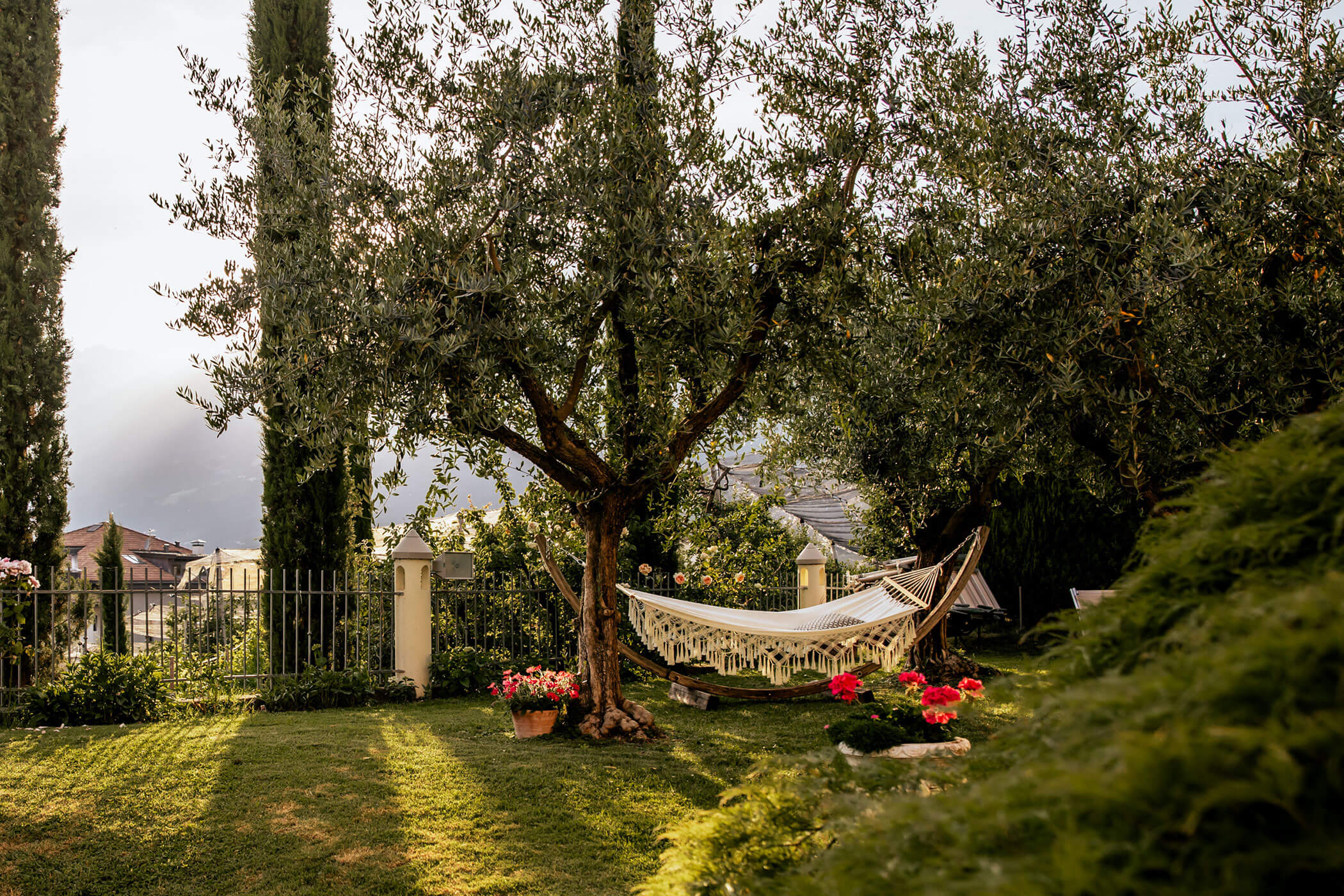 A hammock hangs between two trees in a sunny garden with flowers and a fence in the background. - Hotel Lagrein