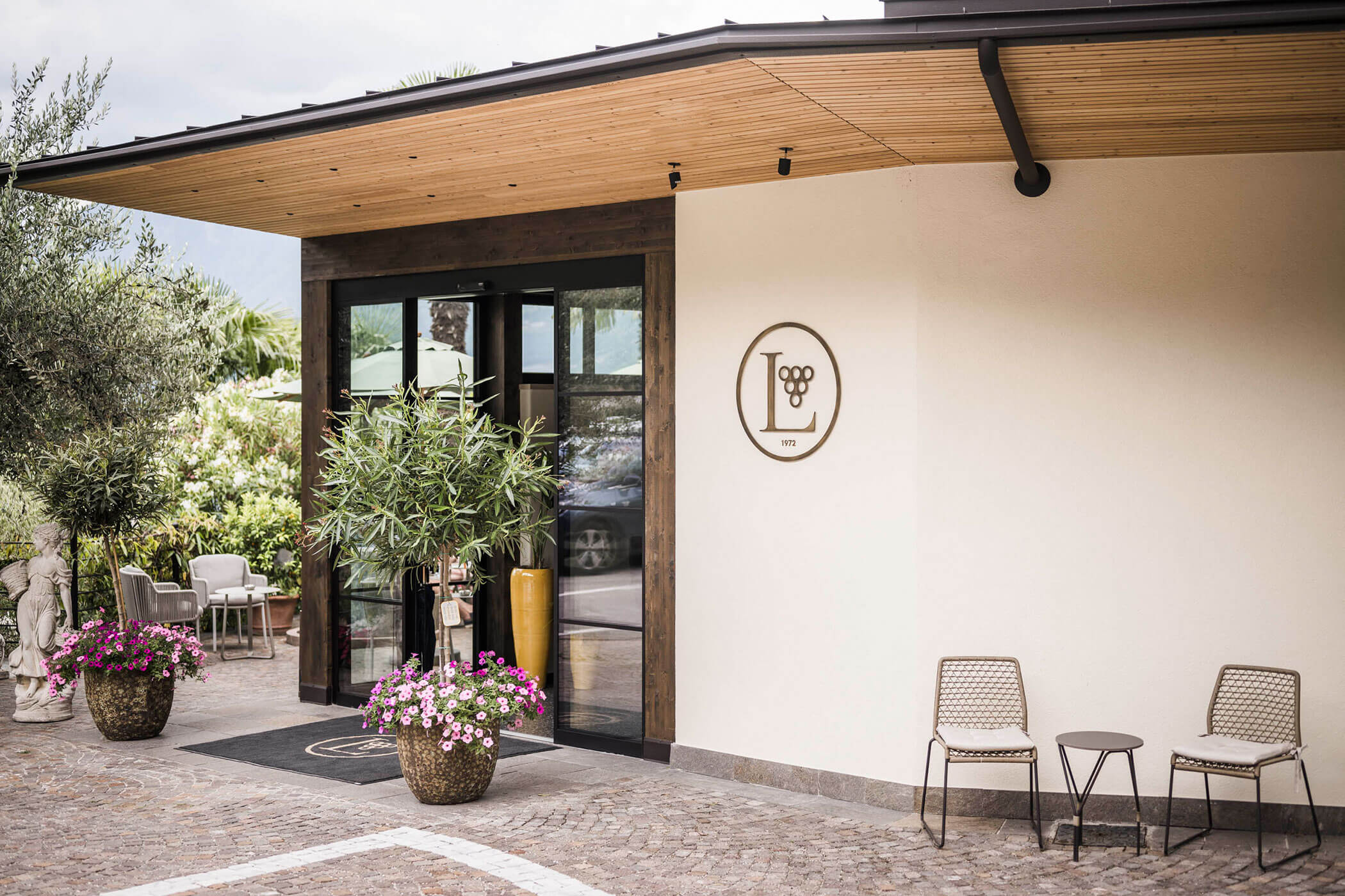 Modern building entrance with glass doors, potted plants, and two chairs by a wall with a circular logo. - Hotel Lagrein
