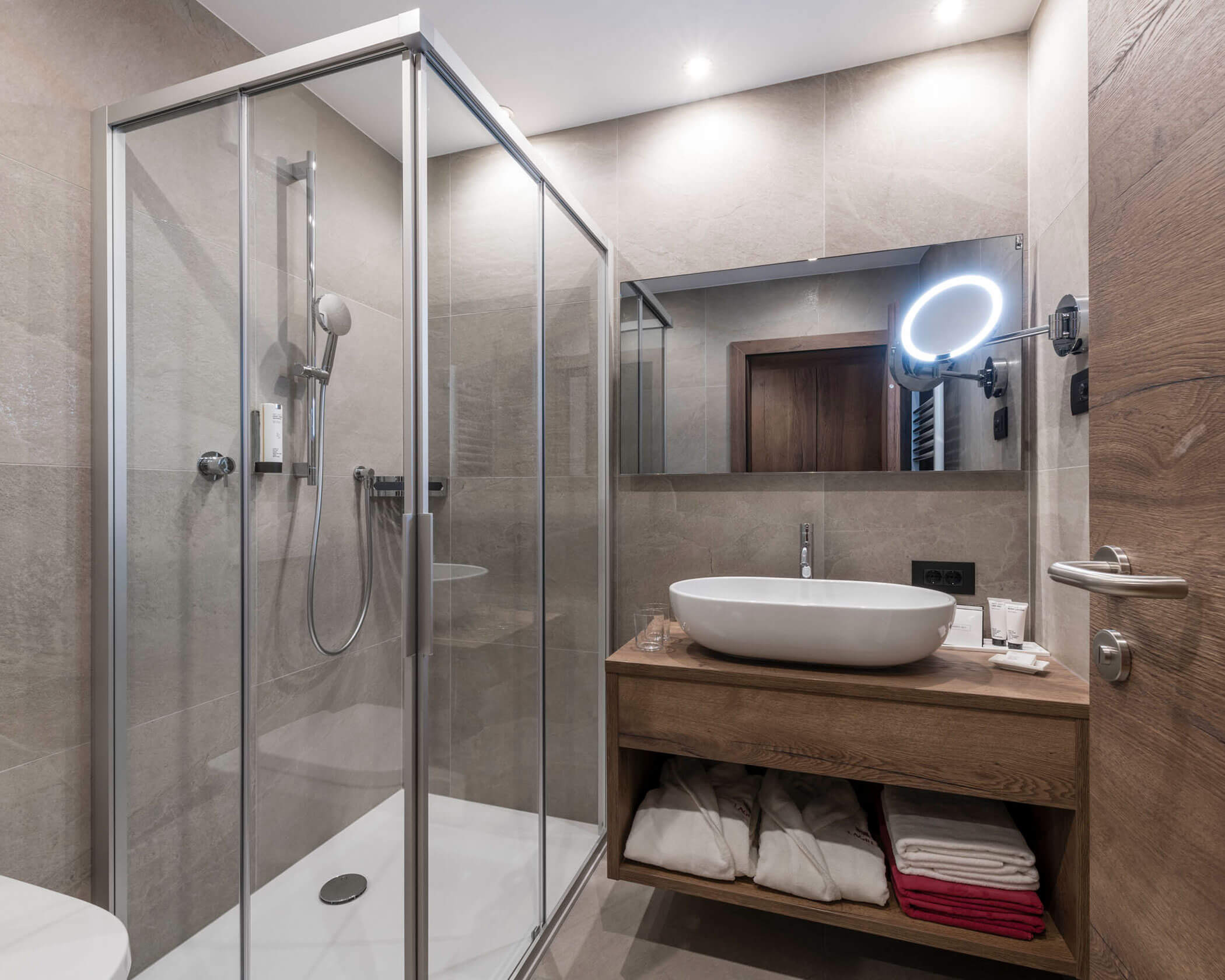 Modern bathroom with a glass shower, vessel sink, wooden vanity, mirror, and folded towels on open shelves. - Hotel Lagrein