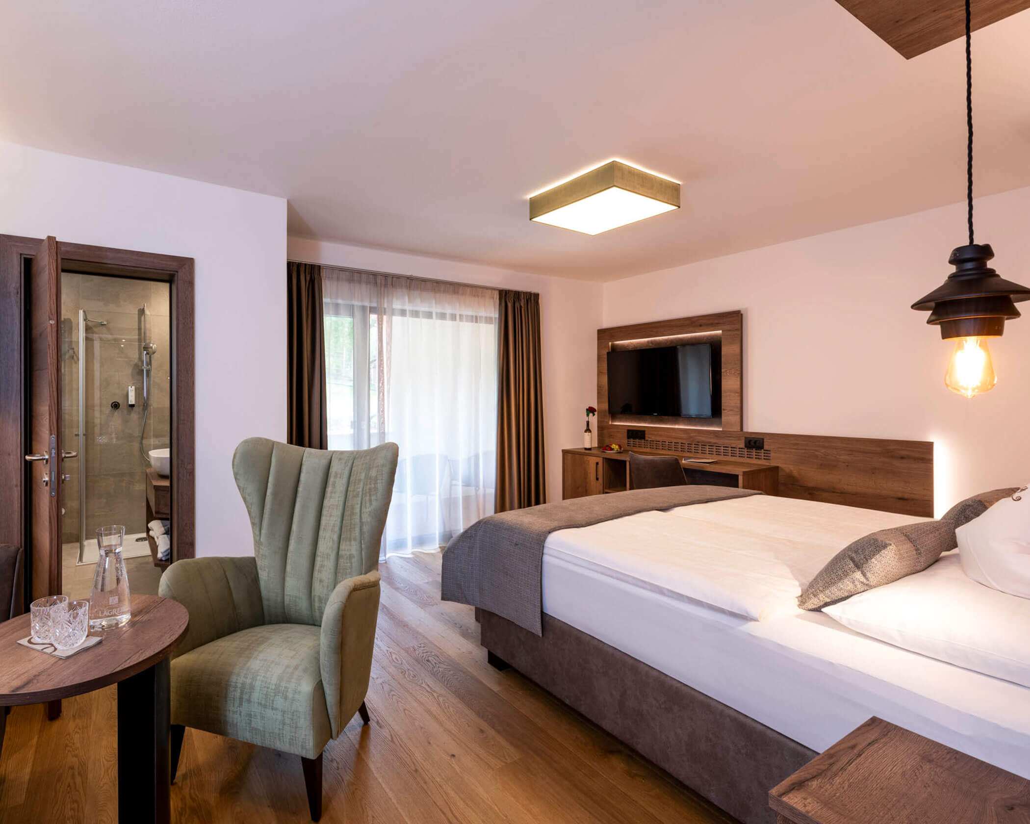 Modern hotel room with a double bed, green armchair, TV, and balcony with sheer curtains. - Hotel Lagrein