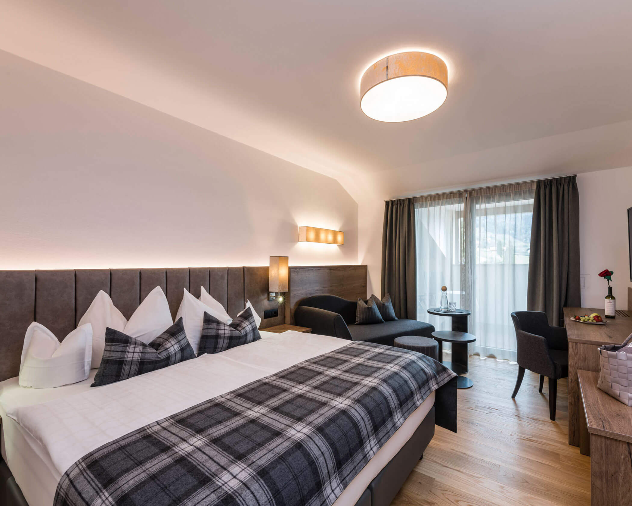 Modern hotel room with a large bed, plaid blanket, sofa, table, chairs, and window with sheer curtains. - Hotel Lagrein